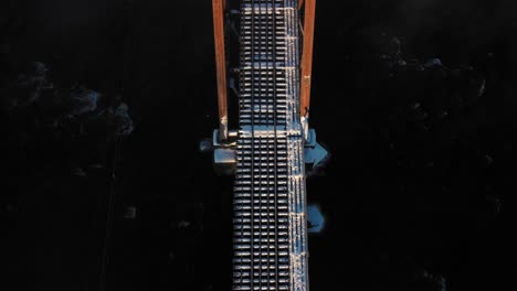 Aerial-TOP-DOWN-flying-over-the-snow-covered-rail-road-trestle-crossing-a-cold-dark-river-at-sunrise