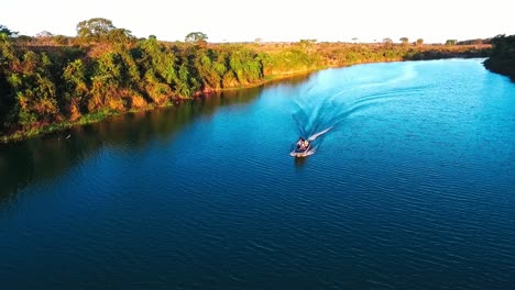A-small-fishing-boat-travels-down-a-deep-blue-river-in-the-evening-sun,-aerial-drone-shot