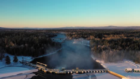Aerial-footage-flying-over-the-dam-on-a-frozen-lake-letting-steaming-water-pour-through-to-the-outlet-river-during-a-winter-sunrise