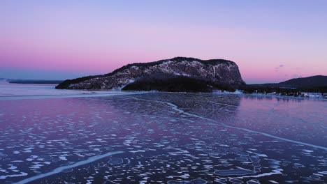 Aerial-ORBIT-around-an-isolated-mountain-in-the-distance-over-a-frozen-lake-at-dawn-with-patterns-of-lines-and-circles-in-the-ice