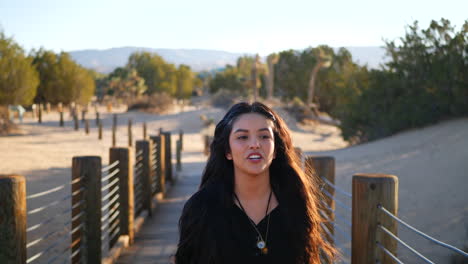 A-pretty-girl-with-long-black-hair-smiling-happily-as-she-walks-along-a-wooden-desert-bridge-in-slow-motion-at-sunset