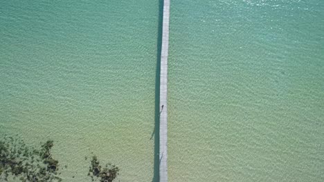 Aerial,-top-down,-birdseye,-drone-shot-over-a-woman-walking-and-a-man-running-on-a-wooden-pier,-while-people-swim-in-the-turquoise-sea,-at-a-paradise-beach,-on-a-sunny-day,-in-Koh-Kood,-Thailand,-Asia