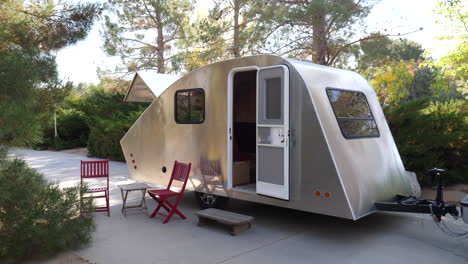 A-new-deluxe-modern-teardrop-travel-trailer-with-chairs-out-for-people-to-enjoy-nature-while-camping-in-the-green-forest-with-trees