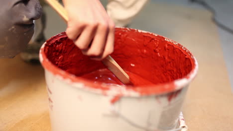 Close-up-of-a-painter-stirring-with-a-wooden-stick-in-a-paint-bucket-full-of-red-paint