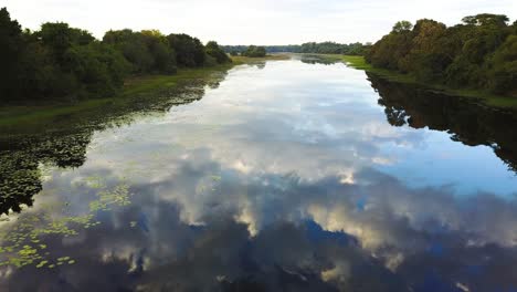 Flying-over-a-calm-river,-as-the-surface-as-a-mirror-reflecting-the-blue-sky-with-clouds