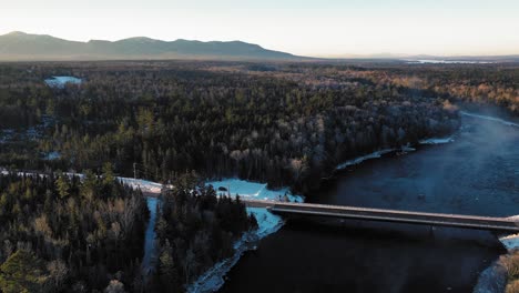Aerial-ORBIT-around-a-bridge-crossing-a-winter-river-revealing-a-mountain-ridge-lit-up-in-the-early-morning-sunshine