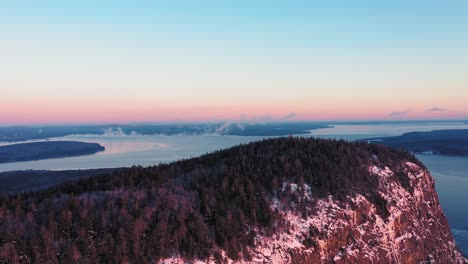 Aerial-view-flying-further-away-from-the-peak-of-a-cliff-faced-mountain-while-seeing-the-misty-freezing-lake-behind-it-during-a-winter-sunrise