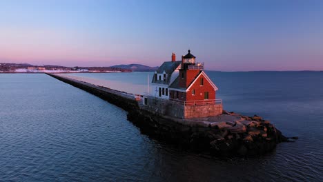 Low-aerial-ORBIT-around-a-red-brick-lighthouse-at-the-end-of-a-snow-dusted-rocky-breakwater-revealing-a-setting-full-moon