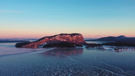 Aerial-view-dropping-down-towards-the-ice-with-a-cliff-faced-mountain-lit-by-a-winter-sunrise