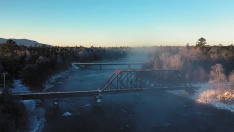 Luftaufnahmen,-Die-Durch-Den-Nebel-Eines-Winterflusses-An-Einem-Eisenbahnbock-Vorbei-Und-Auf-Eine-Brücke-Zufliegen