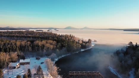 Flying-high-over-the-mist-of-a-curving-winter-river-past-a-railroad-trestle-towards-a-snow-covered-frosted-lake-while-a-truck-drives-along-the-river