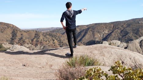 A-young-man-on-a-hike-in-the-California-mountains-stands-at-the-top-of-a-cliff-overlooking-the-landscape-in-slow-motion