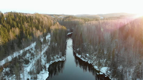 Aerial,-drone-shot,-above-a-river,-sun-flares,-hitting-leafless-trees-and-first-snow-on-the-ground,-on-a-sunny-winter-day,-near-Joensuu,-North-Karelia,-Finland
