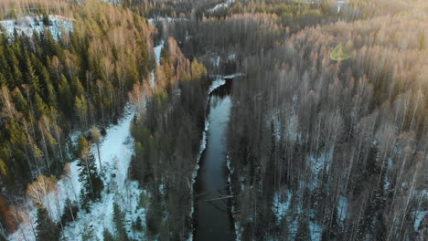 Aerial,-rising,-tilt-up,-drone-shot,-sun-flares,-above-a-river,-surrounded-by-leafless-forest-and-first-snow-on-the-ground,-on-a-sunny-winter-day,-near-Joensuu,-North-Karelia,-Finland