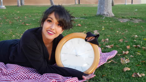 A-pretty-attractive-girl-smiling-with-a-large-clock-to-show-the-time-ticking-away-in-a-park-during-autumn-with-leaves-on-the-ground-and-copy-space