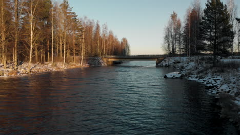 Antenne,-Drohnenschuss,-über-Einem-Fluss,-Auf-Eine-Brücke-Zu,-Umgeben-Von-Blattlosem-Wald-Und-Erstem-Schnee-Auf-Dem-Boden,-An-Einem-Sonnigen-Wintertag,-In-Der-Nähe-Von-Joensuu,-Nordkarelien,-Finnland
