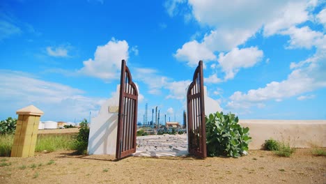 Puertas-De-Hierro-Gigantes-Que-Conducen-A-Un-Cementerio-Vacío-Con-Una-Gran-Refinería-Industrial-En-La-Distancia