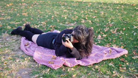 A-young-woman-photographer-taking-pictures-with-her-digital-camera-in-a-park-during-autumn-with-leaves-all-around-the-grass