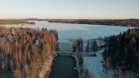 Aerial,-drone-shot,-above-a-river,-over-a-bridge,-towards-a-frozen-lake,-surrounded-by-leafless-forest-and-first-snow-on-the-ground,-on-a-sunny,-autumn-day,-near-Joensuu,-North-Karelia,-Finland