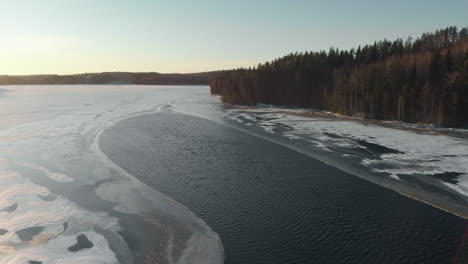 Aerial,-tracking,-drone-shot,-around-first-ice-on-a-lake,-surrounded-by-leafless-forest-and-early-snow-on-the-ground,-on-a-sunny,-autumn-day,-near-Joensuu,-Pohjois-Karjala,-Finland