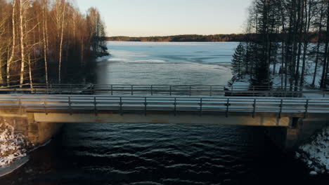Aerial,-reverse,-drone-shot,-above-a-river,-over-a-bridge,-surrounded-by-leafless-forest-and-earley-snow-on-the-ground,-on-a-sunny,-autumn-day,-near-Joensuu,-North-Karelia,-Finland