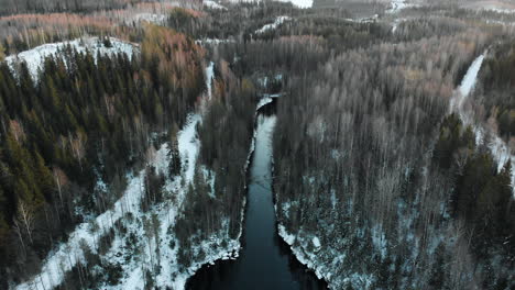 Aerial,-decreasing,-tilt-up,-drone-shot,-sun-flares,-above-a-river,-surrounded-by-leafless-forest-and-first-snow-on-the-ground,-on-a-sunny,-winter-day,-near-Joensuu,-North-Karelia,-Finland
