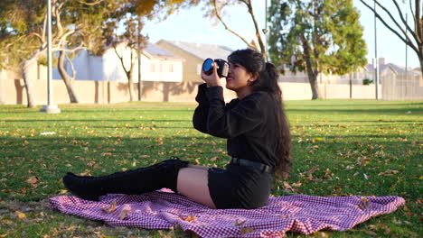 A-hot-girl-taking-pictures-in-slow-motion-with-her-digital-camera-sitting-on-blanket-in-a-park-during-fall-with-autumn-leaves