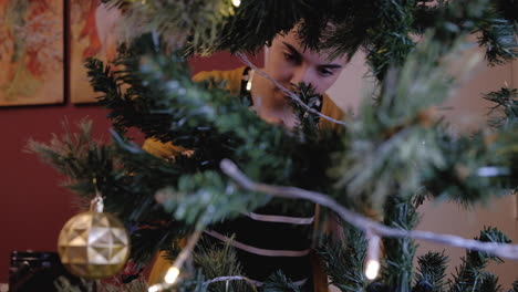 Young-beautiful-caucasian-woman-setting-up-placing-ornaments-on-christmas-tree-framed-between-out-of-focus-branches