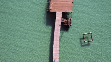 Aerial,-birdseye,-drone-shot,-over-a-asian-woman-walking-on-a-wooden-pier,-surrounded-by-turquoise,-shallow-water,-on-a-sunny-day,-at-Koh-Kood-island,-in-Thailand