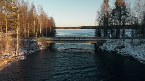Aerial,-drone-shot,-above-a-river,-over-a-bridge,-towards-a-lake,-surrounded-by-leafless-forest-and-first-snow-on-the-ground,-on-a-sunny,-winter-day,-near-Joensuu,-North-Karelia,-Finland