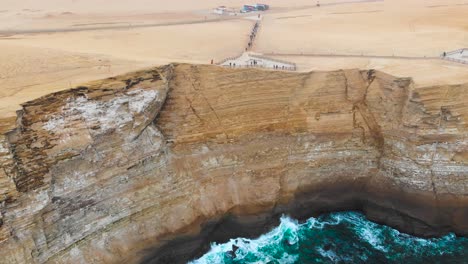 Pull-back-aerial-shot-of-people-standing-at-the-side-of-the-cliff-at-the-ocean-coastline-of-Paracas-National-Park