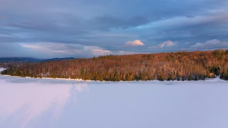 Aerial-footage-flying-up-and-to-the-left-high-above-frozen-lake-at-sunset-with-dark-moody-skies-and-golden-light
