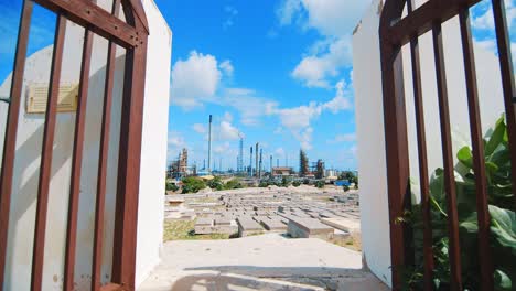 Grandes-Puertas-De-Hierro-Que-Conducen-A-Un-Cementerio-Vacío-En-Curacao-Con-Una-Gran-Refinería-Industrial-Al-Fondo
