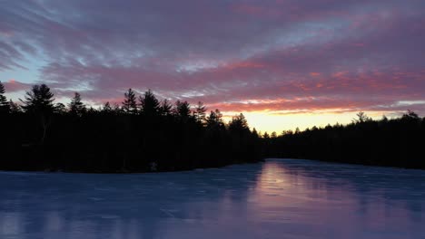 Aerial-SLIDE-over-a-frozen-pond-with-the-forest-silhouetted-against-a-pink-and-yellow-sunrise