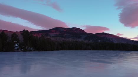 Aerial-footage-flying-low-acrosse-the-surface-of-a-frozen-pond-towards-a-mountain-lit-by-sunrise
