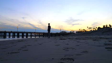 A-man-in-silhouette-alone-on-the-beach-at-Stearns-Wharf-pier-at-sunset-watching-the-ocean-on-the-sand-in-Santa-Barbara,-California