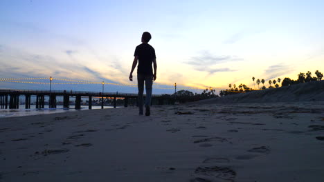 A-man-walks-alone-on-the-beach-to-Stearns-Wharf-pier-at-sunset-watching-ocean-waves-on-the-shore-in-Santa-Barbara,-California