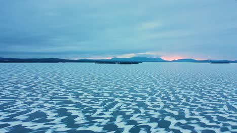 Luftrutsche-über-Einen-Zugefrorenen-See-Mit-Schneebedeckten-Mustern-Im-Eis-Und-Sonnenaufgangsfarbe,-Die-Durch-Die-Bergkette-Am-Horizont-Stößt