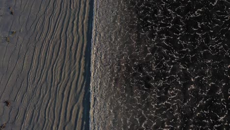 Aerial-TOP-DOWN-flying-over-a-series-of-waves-coming-in-on-a-grey-sand-beach-with-cool-patterns-in-the-sand-and-surf