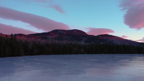 Aerial-SLIDE-flying-low-across-the-surface-of-a-frozen-pond-looking-at-a-mountain-lit-by-sunrise