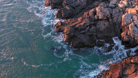 Aerial-TOP-DOWN-of-the-waves-crashing-on-the-rocks-below-Nubble-Lighthouse-in-Maine-lit-by-a-cold-December-sunrise