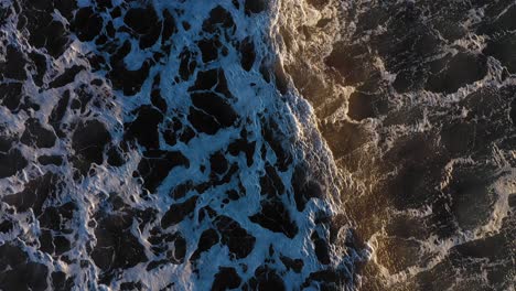 Aerial-TOP-DOWN-flying-slowly-forward-as-a-couple-of-wind-whipped-waves-pass-over-a-dark-ocean-with-cool-patterns-in-the-surf-in-York-Beach-Maine