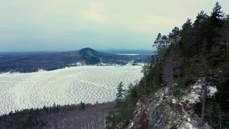 Aerial-footage-flying-close-along-the-ridge-of-a-snowy-mountain-with-a-steep-cliff-face-towards-the-frozen-lake-and-hill-in-the-distance