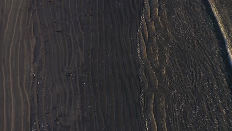 Aerial-TOP-DOWN-slowly-flying-over-a-wave-coming-in-on-a-grey-sand-beach-with-cool-patterns-in-the-sand-and-surf