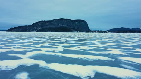 Aerial-footage-flying-low-over-a-frozen-lake-with-snowy-patterns-towards-an-isolated-mountain-with-a-steep-cliff