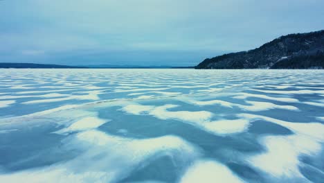 Aerial-footage-flying-low-over-a-frozen-lake-with-snowy-patterns-towards-the-edge-of-a-cliff-rising-out-of-the-ice