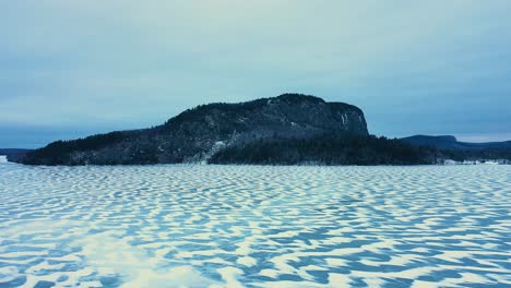 Aerial-SLIDE-over-a-frozen-lake-with-snowy-patterns-past-an-isolated-mountain-with-a-steep-cliff-face