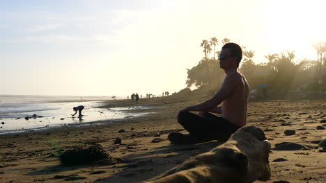 Ein-Junger-Mann-Reisender-Auf-Dem-Sand-Von-Rincon-Point-Beach-In-Kalifornien,-Der-Die-Surfer-Und-Wellen-Bei-Sonnenuntergang-Mit-Palmen-Beobachtet