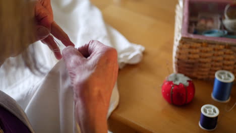 The-hands-of-an-aging-elderly-woman-sewing-a-button-on-a-white-dress-shirt-by-hand-with-a-pincushion-and-spools-of-thread
