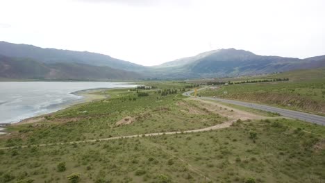 Aerial-travelling-between-reservoir-waters-road-with-views-ahead-of-Mountain-range,-El-Mollar,-Dique-la-Angostura